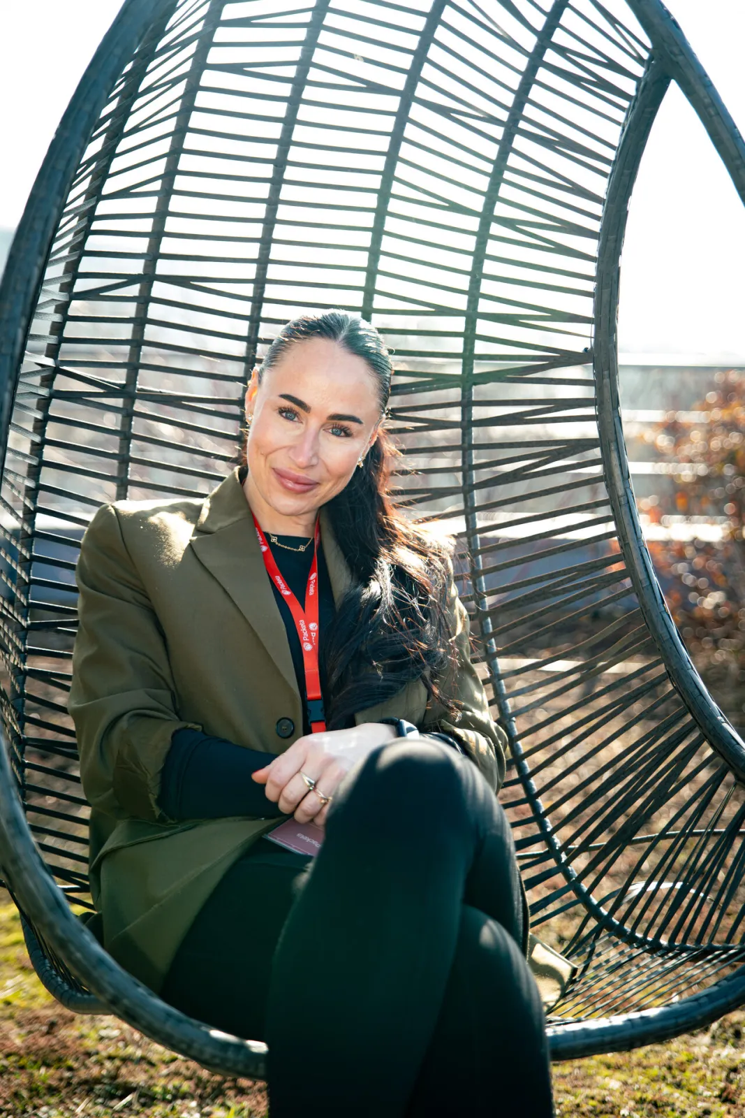 Caroline S. Karda, Group Head of Talent Acquisition ve společnosti Packeta, sedí venku v černém proutěném závěsném křesle a usmívá se do objektivu. Na sobě má tmavě zelené sako a na krku červenou šňůrku Packeta. Foceno pro sérii HR Vibecheck od agentury VERYGUD.