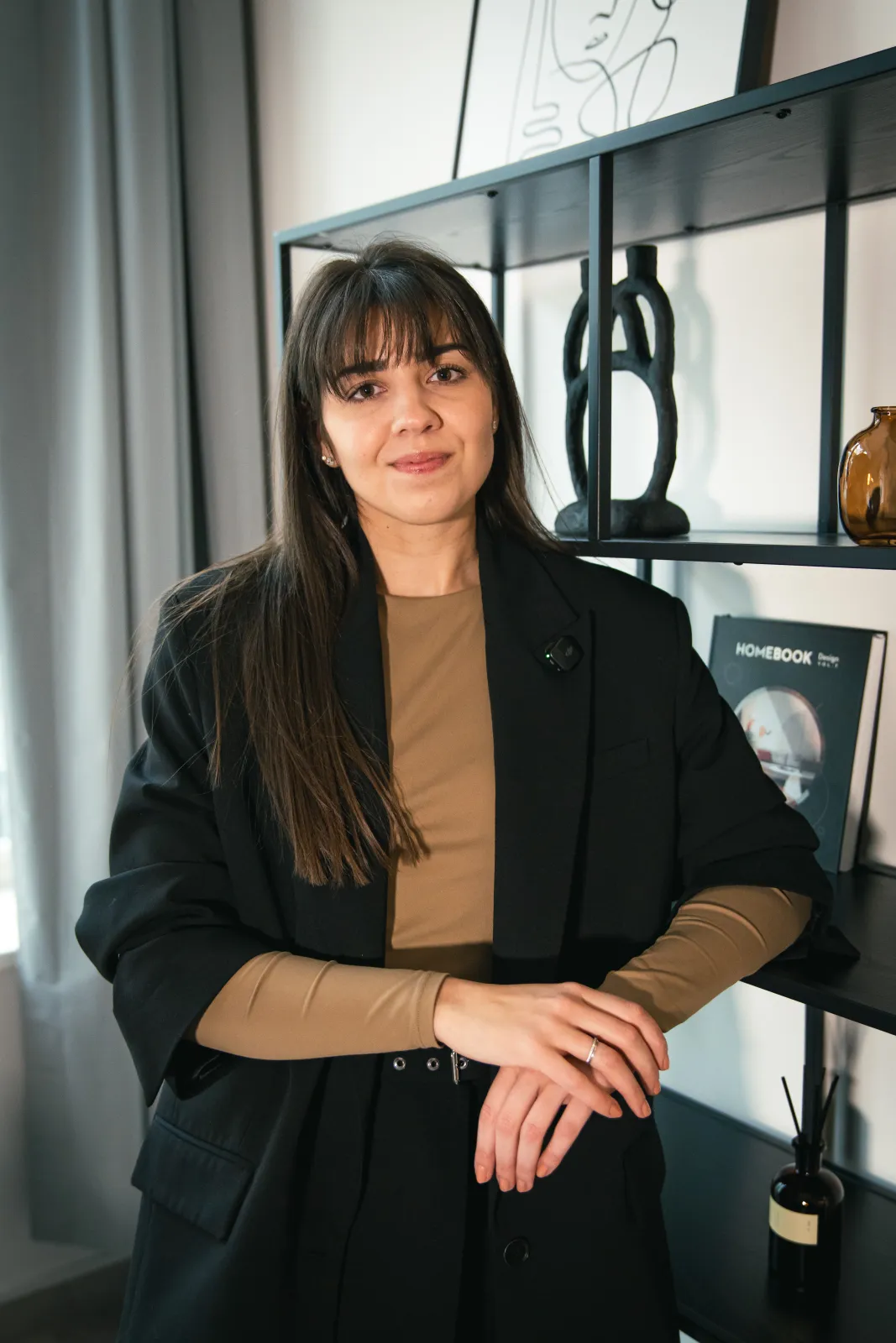 Denisa Fousová, Head of HR ve společnosti Bonami, se jemně usmívá do objektivu během natáčení série HR Vibecheck od agentury VERYGUD. Na sobě má hnědé triko s dlouhým rukávem a černé sako, na kterém má připnutý malý klopový mikrofon. Stojí v moderních kancelářích Bonami a ležérně se opírá o černý kovový regál s designovými dekoracemi a knihou.